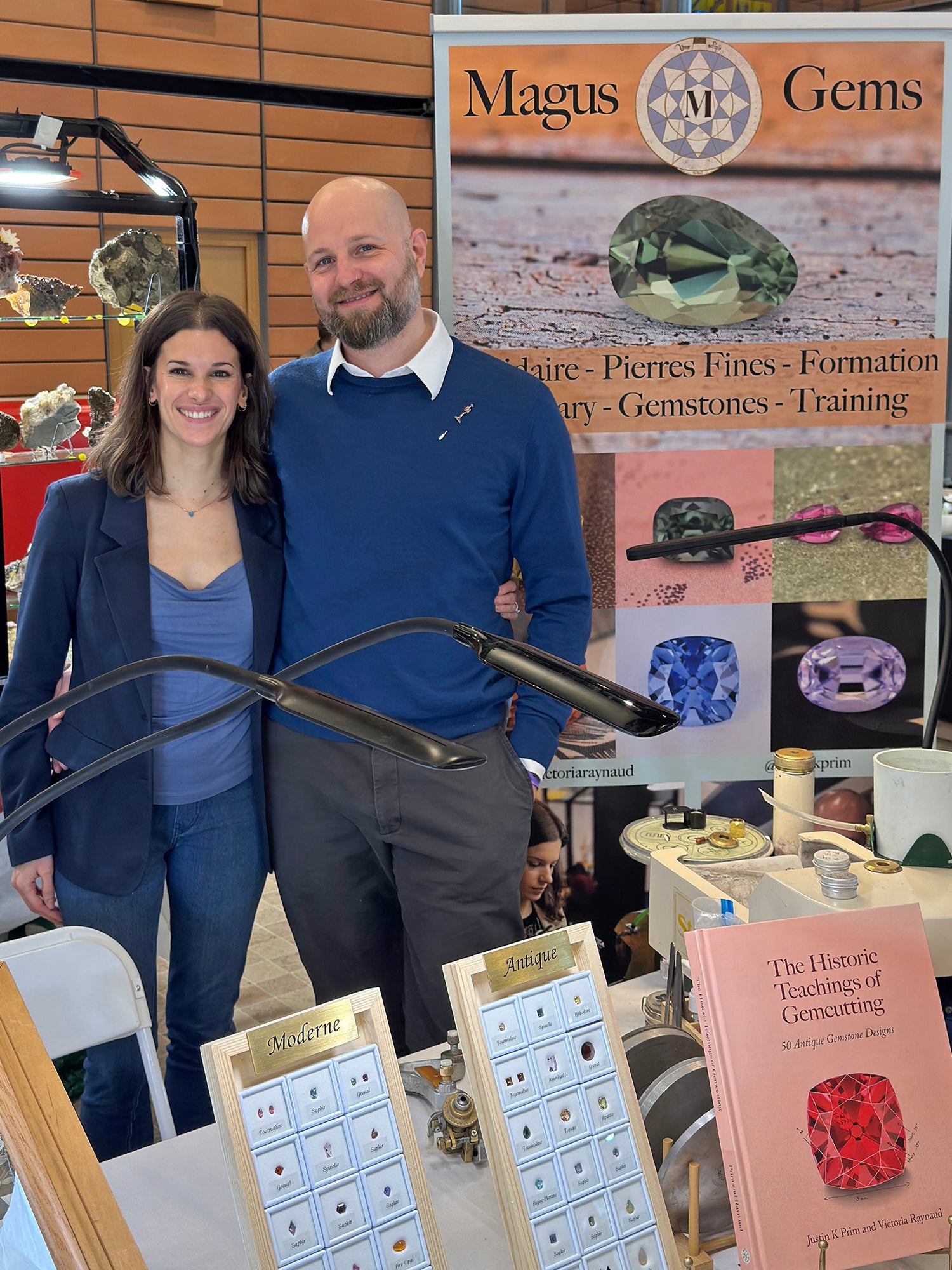 A man and a woman stand behind a display table at a gemstone exhibition, with jewelry pieces, cards, and a 'Magus Gems' banner visible in the background.